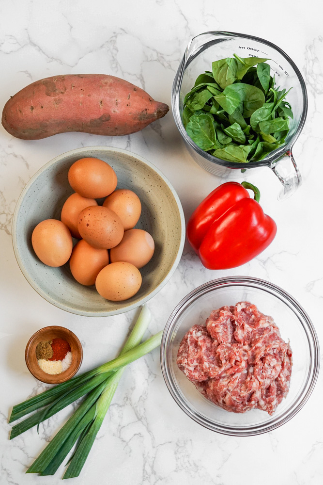 Ingredients laid out on marble include sweet potato, spinach, eggs, bell pepper, sausage, green onion and spices